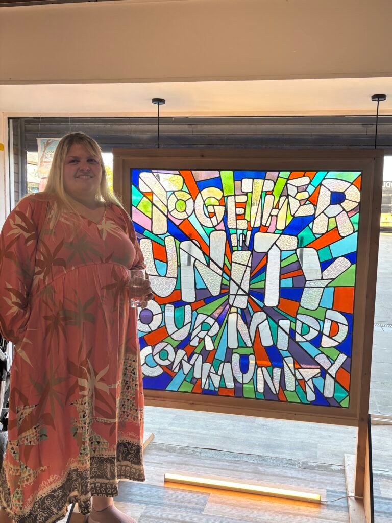 Laura standing next to a stained glass window 'Together' which has the phrase 'Together in Unity, Our Midd Community' in multicoloured glass panels.